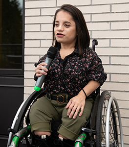 Une femme atteinte de nanisme tenant un microphone, assise dans un fauteuil roulant (c) Disabled and Here