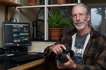 James Smedley - smiling and holding a camera sitting in front of a computer